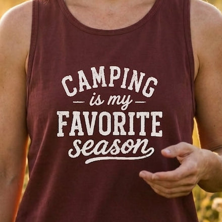 A woman standing in a sunlit grassy field wearing a sleeveless maroon tank top with the white printed text 'CAMPING is my FAVORITE season'.