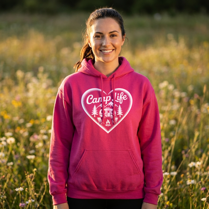 Smiling woman in a bright pink Camp Life heart hoodie standing in a wildflower field at sunset