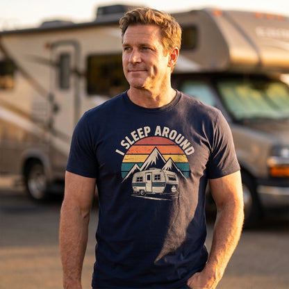 A man wearing a navy blue graphic t-shirt with a retro mountain and camper design that reads 'I SLEEP AROUND'. He is standing outdoors in front of a blurred recreational vehicle at sunset.