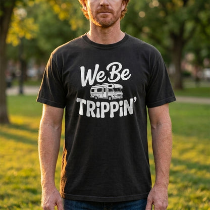 A man wearing a black graphic t-shirt with a white 'We Be Trippin'' and RV design, standing outdoors in a park setting.