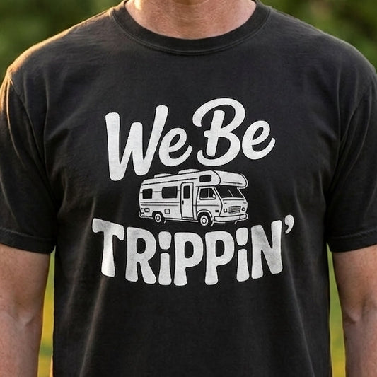 A man wearing a black graphic t-shirt with a white 'We Be Trippin'' and RV design, standing outdoors in a park setting.
