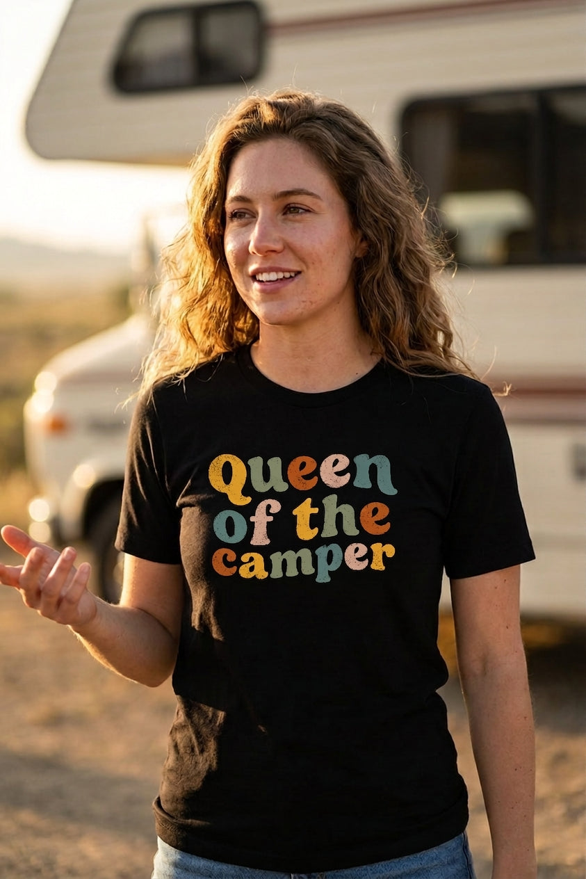 A woman wearing a black t-shirt with the text 'Queen of the camper' in a retro, multi-colored font, standing outdoors in front of a blurred recreational vehicle.