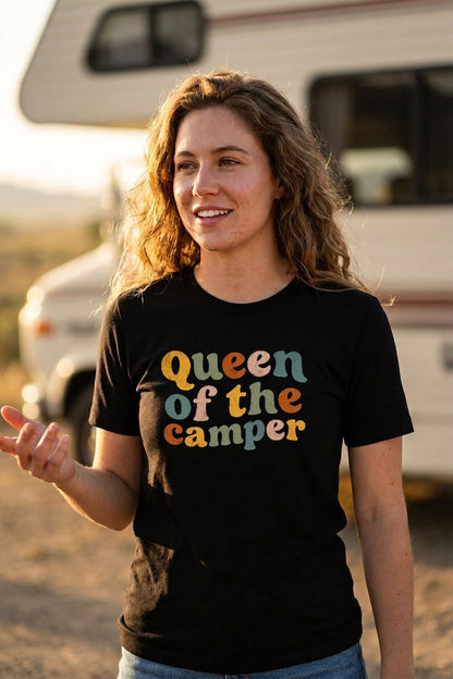 A woman wearing a black t-shirt with the text 'Queen of the camper' in a retro, multi-colored font, standing outdoors in front of a blurred recreational vehicle.