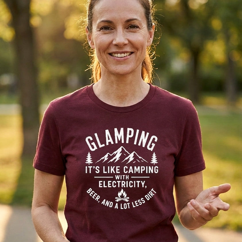 A woman smiling outdoors wearing a maroon short-sleeve t-shirt with a white graphic print that reads 'GLAMPING IT'S LIKE CAMPING WITH ELECTRICITY' and 'BEER AND A LOT LESS DRINK'.