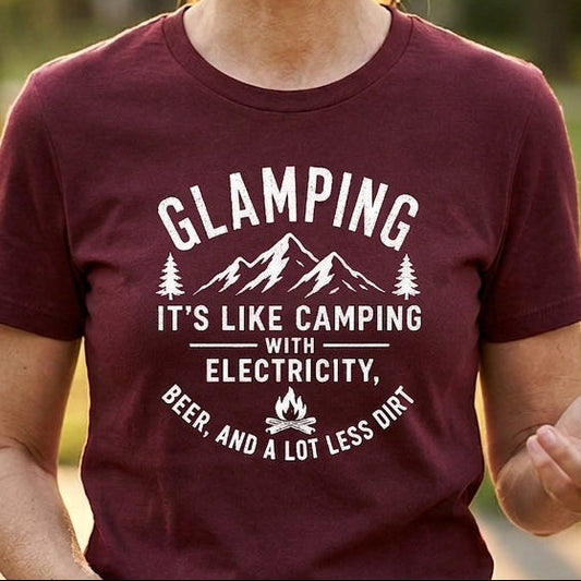 A woman smiling outdoors wearing a maroon short-sleeve t-shirt with a white graphic print that reads 'GLAMPING IT'S LIKE CAMPING WITH ELECTRICITY' and 'BEER AND A LOT LESS DRINK'.