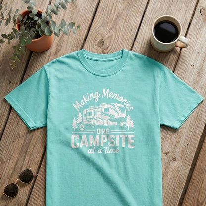 A mint green short-sleeve t-shirt with a white graphic print reading 'Making Memories One Campsite at a Time' with a camper illustration, laid flat on a rustic wooden surface with a mug, book, sunglasses, and plant nearby.