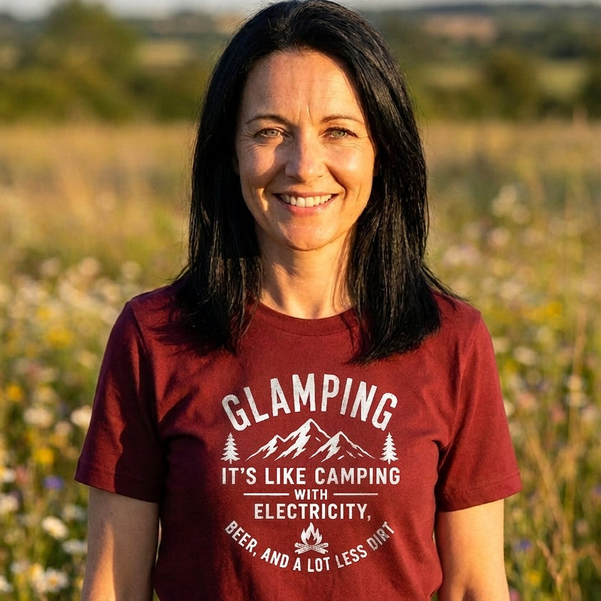 A smiling woman with dark hair stands outdoors in a sunlit field wearing a maroon short-sleeve graphic T-shirt with white text that reads, 'Glamping it's like camping with electricity, beer, and a lot less sleep.'.