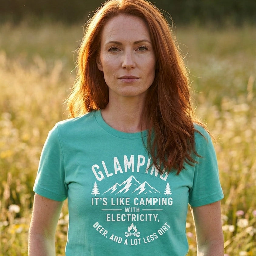 A woman with red hair stands in a grassy field at sunset wearing a teal t-shirt with a white graphic that reads 'GLAMPING IT'S LIKE CAMPING WITH ELECTRICITY, BEER, AND A LOT LESS DIRT'.