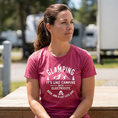 A woman sitting outdoors at a wooden table wearing a pink graphic t-shirt that reads 'GLAMPING IT'S LIKE CAMPING WITHOUT ELECTRICITY, EVER. AND A LOT LESS SHIT.