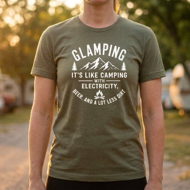 A person standing outdoors at sunset wearing a military green graphic t-shirt with a white printed design that reads 'GLAMPING IT'S LIKE CAMPING WITH ELECTRICITY' and 'BEER AND A LOT LESS DRINK'.