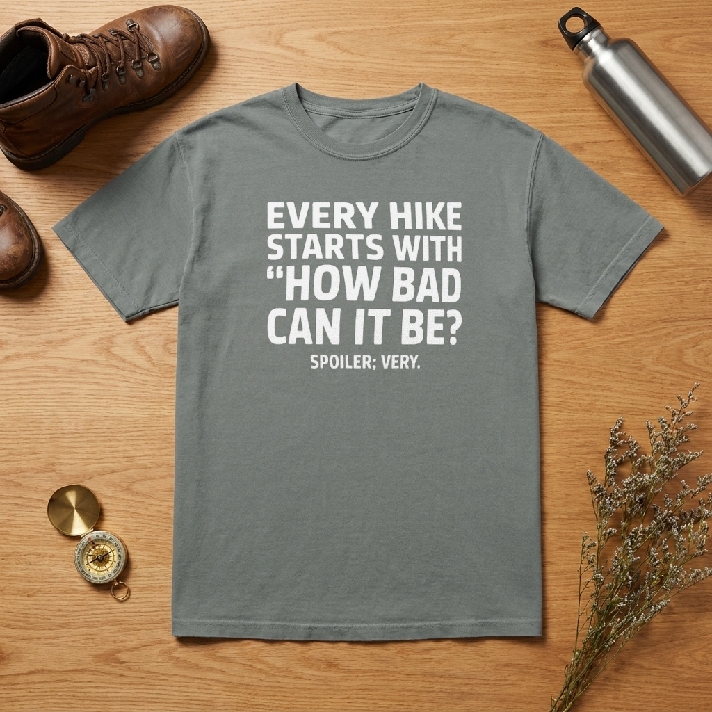 A gray short-sleeve graphic T-shirt laid flat on a wooden surface, surrounded by hiking-themed props including a brown boot, metal water bottle, compass, dried flowers, and a small round accessory. The shirt has large white text reading, 'EVERY HIKE STARTS WITH "HOW BAD CAN IT BE?" SPOILER: VERY.