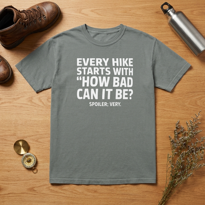 A gray short-sleeve graphic T-shirt laid flat on a wooden surface, surrounded by hiking-themed props including a brown boot, metal water bottle, compass, dried flowers, and a small round accessory. The shirt has large white text reading, 'EVERY HIKE STARTS WITH "HOW BAD CAN IT BE?" SPOILER: VERY.