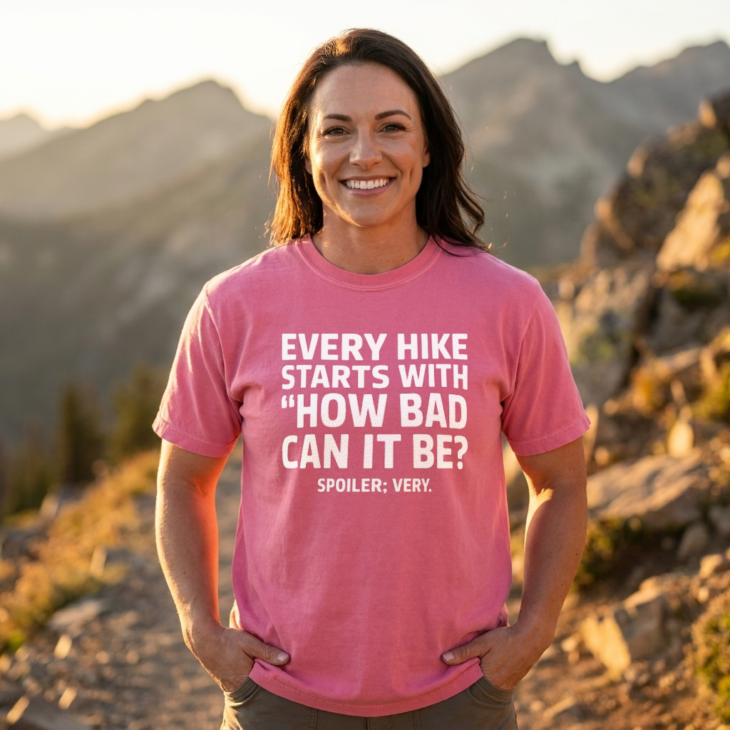 A smiling woman wearing a pink graphic t-shirt stands outdoors in a mountain setting. The t-shirt features the white text 'EVERY HIKE STARTS WITH "HOW BAD CAN IT BE?" SPOILER: VERY.