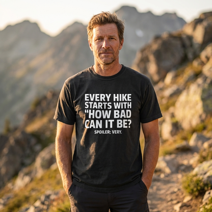 A man standing outdoors on a rocky mountain trail wearing a black graphic t-shirt with white text that reads 'EVERY HIKE STARTS WITH "HOW BAD CAN IT BE?" SPOILER: VERY.'.