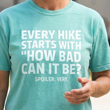 A woman wearing a teal graphic t-shirt with white text that reads 'EVERY HIKE STARTS WITH "HOW BAD CAN IT BE?" SPOILER: VERY.' She is standing outdoors in front of a blurred travel trailer and trees.
