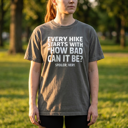 A woman wearing a dark gray graphic t-shirt with white text that reads 'EVERY HIKE STARTS WITH "HOW BAD CAN IT BE?" SPOILER: VERY.' standing in a grassy park.