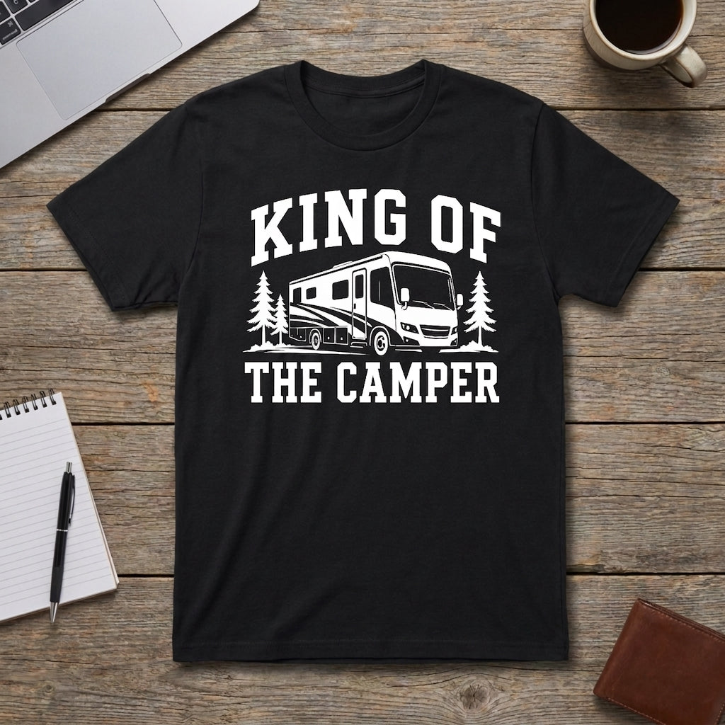 Black King of the Camper t-shirt with a white RV and pine trees graphic laid flat on a wooden table with a laptop, coffee mug, notebook, pen, and wallet.