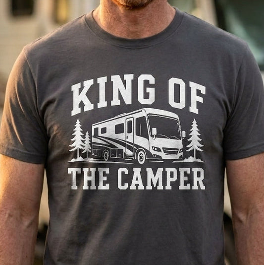 A smiling man with red hair and a beard stands outdoors in front of an RV, wearing a dark gray graphic t-shirt that reads 'KING OF THE CAMPER' with a camper van and pine tree illustration.