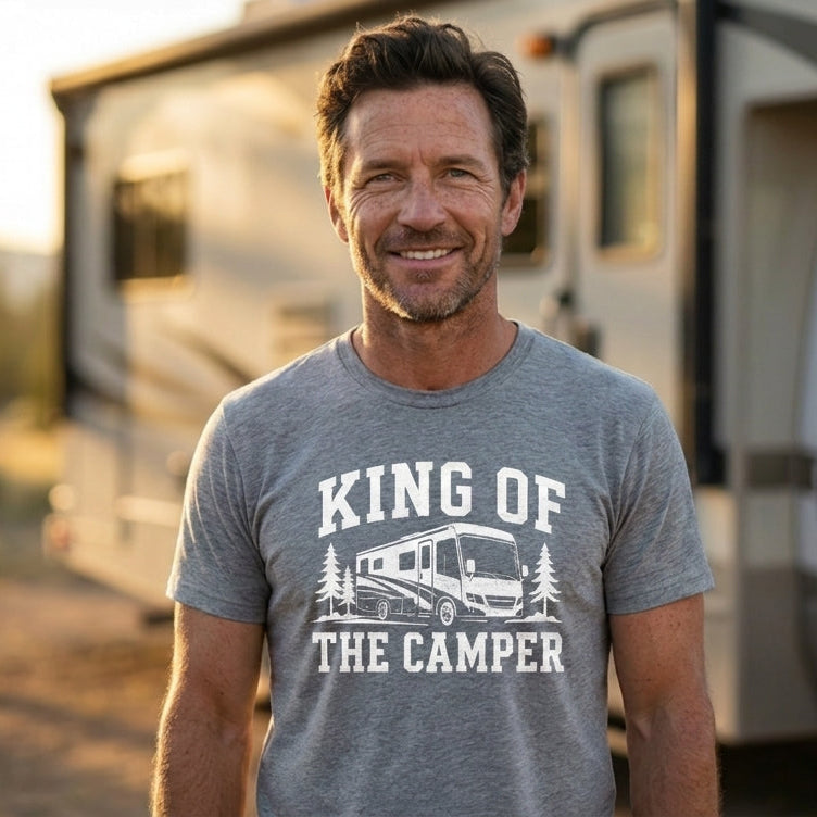 Smiling man in a heather gray King of the Camper t-shirt with a white graphic, standing in front of a white RV at golden hour.