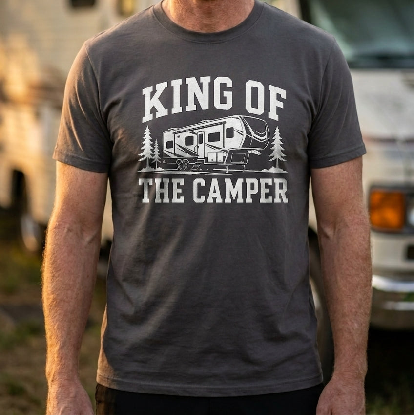 A smiling man with red hair and a beard stands outdoors in front of an RV, wearing a dark gray graphic t-shirt that reads 'KING OF THE CAMPER' with a camper illustration.