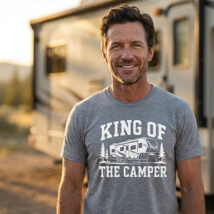 A smiling man wearing a heather gray graphic t-shirt that reads 'KING OF THE CAMPER' with a camper illustration, standing outdoors in front of an RV during golden hour.