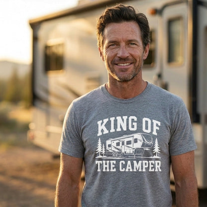 A smiling man wearing a heather gray graphic t-shirt that reads 'KING OF THE CAMPER' with a camper illustration, standing outdoors in front of an RV during golden hour.