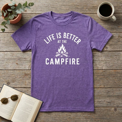 A heather purple short-sleeve t-shirt with a white graphic reading 'LIFE IS BETTER AT THE CAMPFIRE' and a campfire illustration, laid flat on a rustic wooden surface with a book, sunglasses, plant, and coffee mug as props.