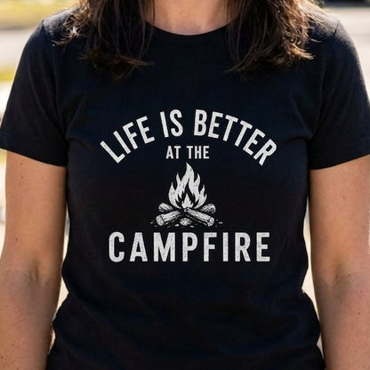 A smiling woman stands outdoors at a campground or RV park wearing a black short-sleeve T-shirt with the white text 'LIFE IS BETTER AT THE CAMPFIRE' and a small campfire graphic on the front.