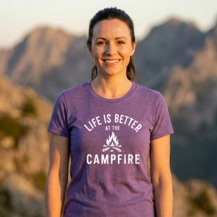 A woman wearing a purple graphic t-shirt with the text 'LIFE IS BETTER AT THE CAMPFIRE' stands in front of a blurred mountain landscape at sunset.