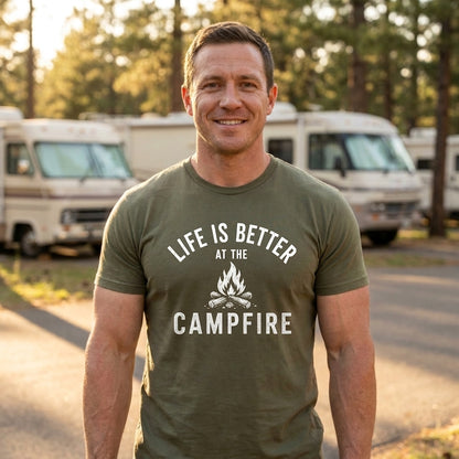 A smiling man wearing an olive green t-shirt with a white graphic that reads 'LIFE IS BETTER AT THE CAMPFIRE' stands outdoors in front of campervans and trees.