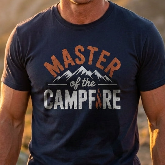Smiling man in a navy Master of the Campfire t-shirt with orange, white, and gray lettering, standing in front of mountains at sunset.
