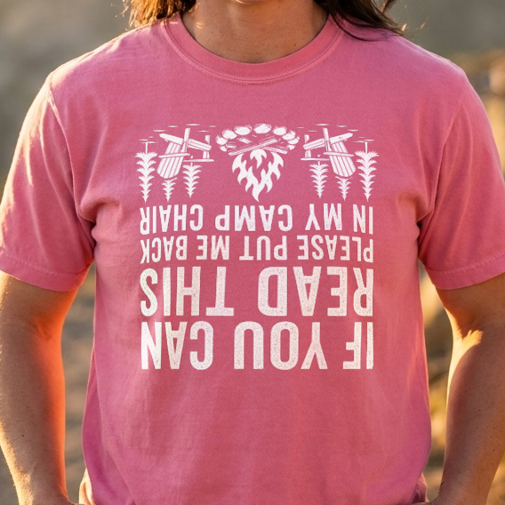 Close-up of a person in a pink If You Can Read This camp chair t-shirt with upside-down white text and tree icons, outdoors at golden hour.