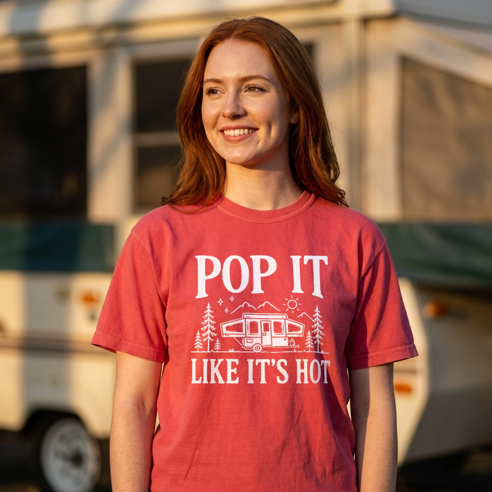 A smiling young woman with red hair stands outdoors in front of a blurred RV, wearing a faded red crew-neck t-shirt with a white graphic that reads 'POP IT LIKE IT'S HOT' with a camper illustration.