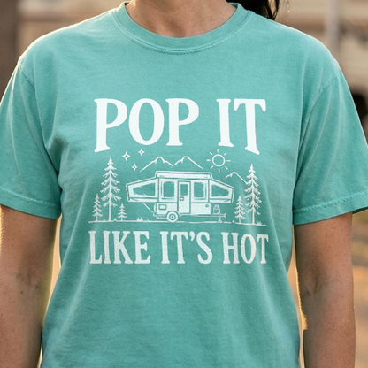 A woman wearing a mint green graphic t-shirt with a white print that reads 'POP IT LIKE IT'S HOT' and features a camper trailer design, standing outdoors in front of a blurred background of RVs and trees.