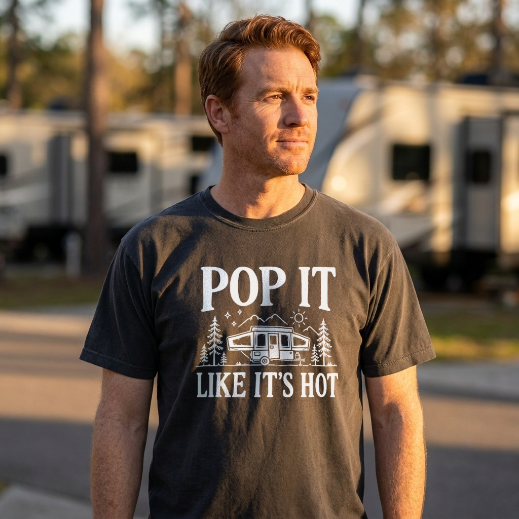 A man wearing a dark charcoal gray t-shirt with a white graphic print that reads 'POP IT LIKE IT'S HOT' with a camper illustration, standing outdoors in front of blurred RVs.