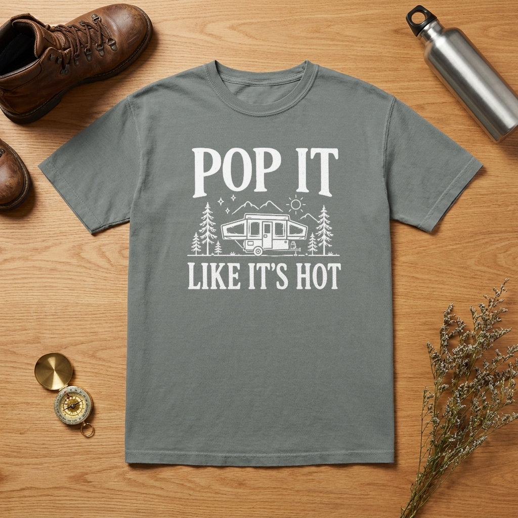 A gray short-sleeve t-shirt with a white graphic print reading 'POP IT LIKE IT'S HOT' and showing a camping trailer, trees, and mountains. The shirt is laid flat on a wooden surface surrounded by lifestyle props including a brown hiking boot, a silver water bottle, a compass, and dried flowers.