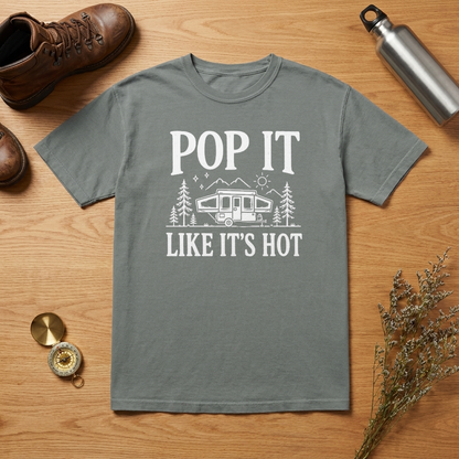A gray short-sleeve t-shirt with a white graphic print reading 'POP IT LIKE IT'S HOT' and showing a camping trailer, trees, and mountains. The shirt is laid flat on a wooden surface surrounded by lifestyle props including a brown hiking boot, a silver water bottle, a compass, and dried flowers.