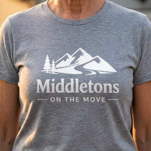 A smiling older woman stands outdoors in front of a large recreational vehicle, wearing a heather gray T-shirt with a white mountain graphic and the text 'Middletons ON THE MOVE' on the front.