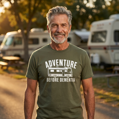 A smiling older man standing outdoors at a campsite wearing an olive green t-shirt with a white graphic that reads 'ADVENTURE BEFORE DEMENTIA' with a bus illustration.