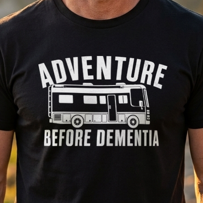 A smiling man standing outdoors on a mountain trail at sunset, wearing a black graphic T-shirt with the words “ADVENTURE BEFORE DEMENTIA” and a bus illustration printed in white.