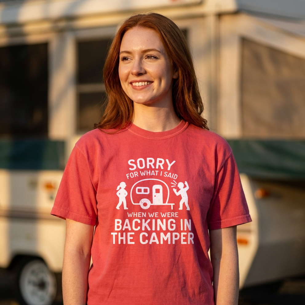 A smiling young woman with red hair stands outdoors in front of a blurred RV, wearing a coral-red graphic T-shirt with white text that reads, 'SORRY FOR WHAT I SAID WHEN WE WERE BACKING IN THE CAMPER.'.