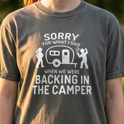 A woman wearing a dark gray graphic t-shirt in an outdoor park setting. The t-shirt features a white graphic of a camper and the text 'SORRY FOR WHAT I SAID WHEN WE WERE BACKING IN THE CAMPER'.