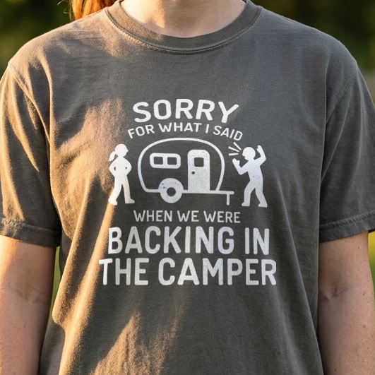 A woman wearing a dark gray graphic t-shirt in an outdoor park setting. The t-shirt features a white graphic of a camper and the text 'SORRY FOR WHAT I SAID WHEN WE WERE BACKING IN THE CAMPER'.