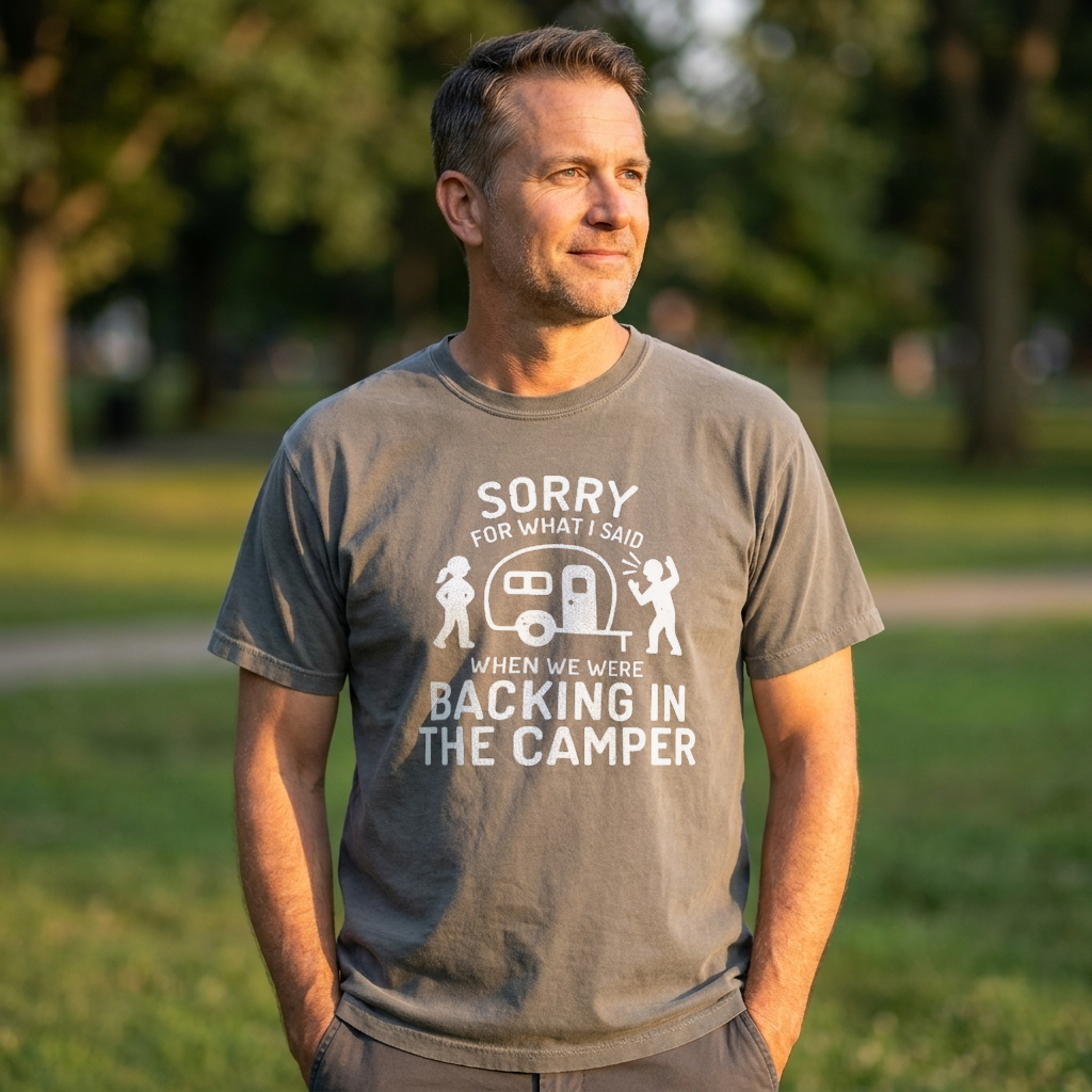 A man wearing a gray graphic t-shirt with a white print that reads 'SORRY FOR WHAT I SAID WHEN WE WERE BACKING IN THE CAMPER' with a camper illustration, standing in a park.