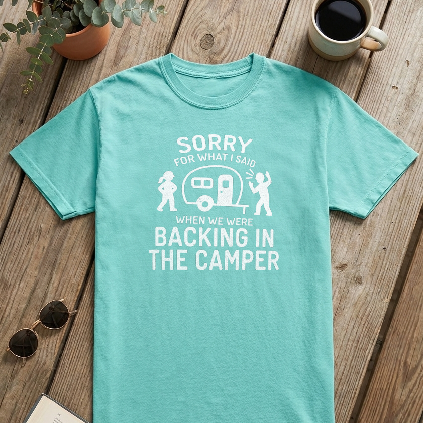 A mint green graphic t-shirt laid flat on a rustic wooden table, surrounded by lifestyle props including an open book, sunglasses, a potted plant, a coffee mug, and a camper-themed print on the front.
