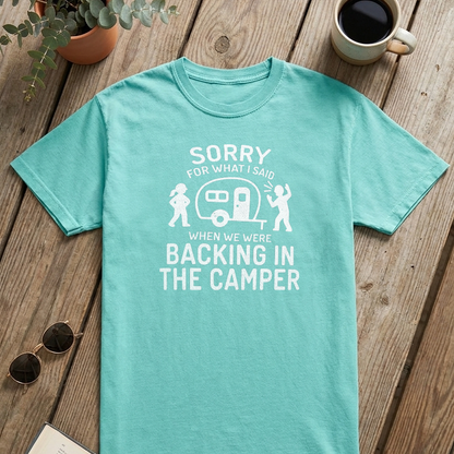 A mint green graphic t-shirt laid flat on a rustic wooden table, surrounded by lifestyle props including an open book, sunglasses, a potted plant, a coffee mug, and a camper-themed print on the front.