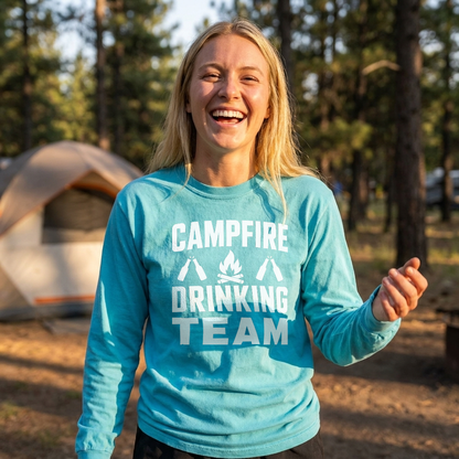 Laughing woman in a teal Campfire Drinking Team long sleeve shirt standing near a tent in the pines