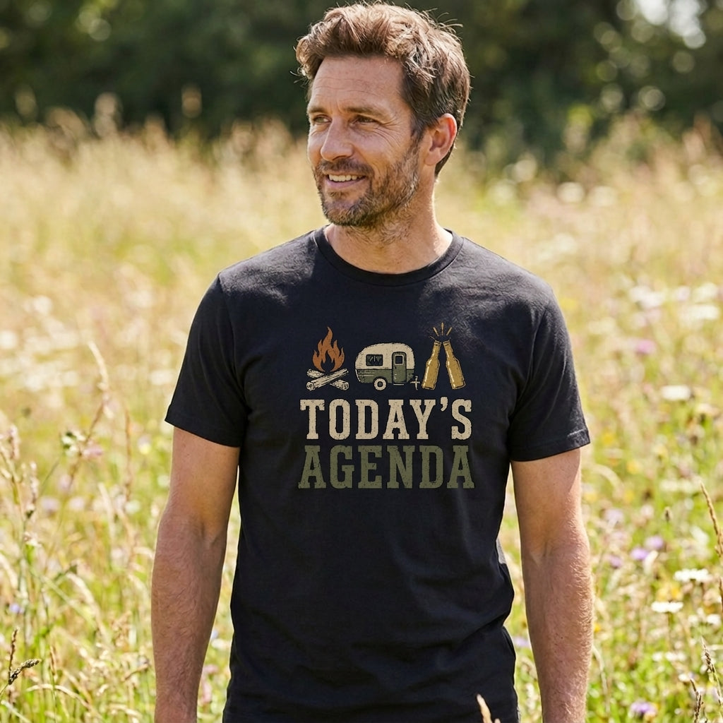 A man wearing a black graphic t-shirt in an outdoor grassy field. The t-shirt features a graphic of a campfire, a vintage camper van, a tent, and the text 'TODAY'S AGENDA' in a distressed font.