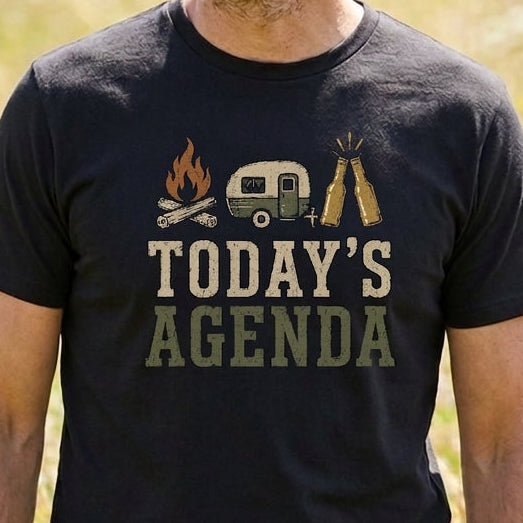 A man wearing a black graphic t-shirt in an outdoor grassy field. The t-shirt features a graphic of a campfire, a vintage camper van, a tent, and the text 'TODAY'S AGENDA' in a distressed font.