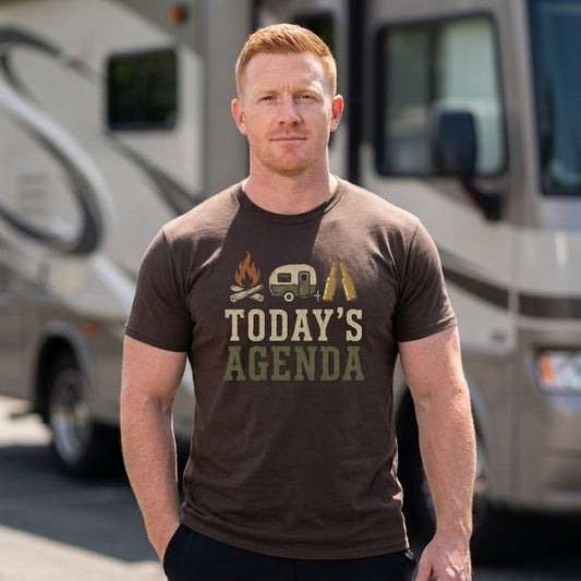 A man wearing a brown graphic t-shirt with 'TODAY'S AGENDA' and camping-themed illustrations, standing in front of a blurred recreational vehicle.